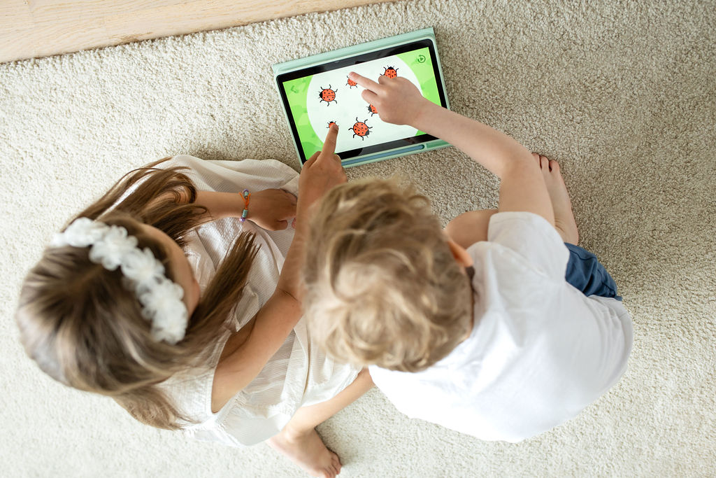 Dziewczynka i chłopiec siedzą razem na dywanie i wspólnie grają w edukacyjną grę na tablecie, licząc biedronki na ekranie.