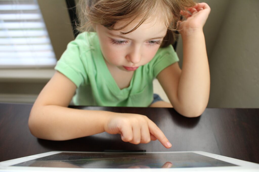 Menina pequena usando o dedo para tocar na tela de um tablet apoiado sobre uma mesa.