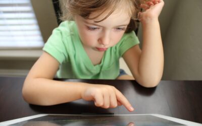 Menina pequena usando o dedo para tocar na tela de um tablet apoiado sobre uma mesa.