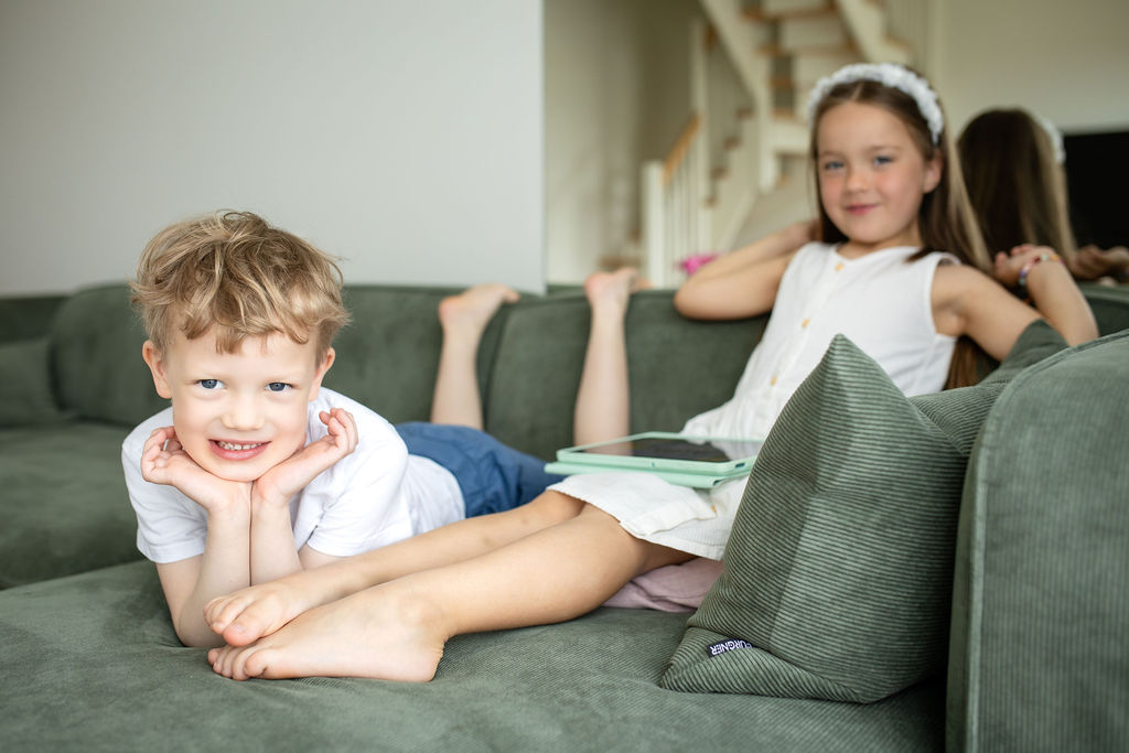 Smaidošs zēns un meitene atpūšas kopā uz dīvāna, meitenei rokās planšetdators ar ALPA Kids lietotni.